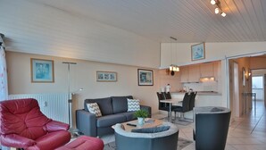 Ett modernt vardagsrum med en röd läderfåtölj, en grå soffa och ett soffbord med en växt. Det finns en matplats med ett bord och stolar, och ett kök syns i bakgrunden.