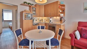 A compact kitchen with wooden cabinets, a microwave, and a dining area with a round table and chairs. - Strandburg Appartement 11 (Scharbeutz)
