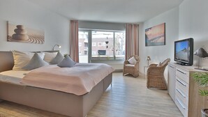 A modern bedroom with a large bed, a television, a wicker chair, and a view of the city through the window. - Barke Appartement 202 (Scharbeutz)