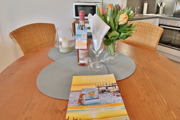 A dining table with a placemat, a bottle of wine, a glass, a vase with flowers, and a welcome brochure. - Barke Appartement 202 (Scharbeutz)