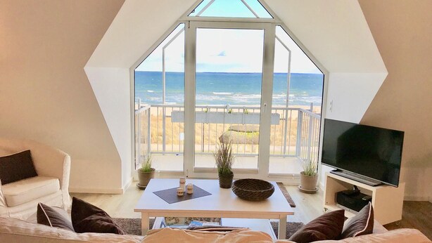 Ein Wohnzimmer mit großem Fenster zum Strand, einem Flachbildfernseher und einer gemütlichen Sitzecke.