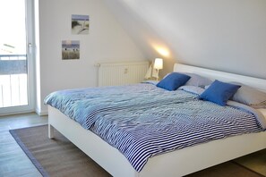 A bedroom with a bed, pillows, a nightstand, a lamp, and a window. - Stranddüne Appartement 12 (Haffkrug)