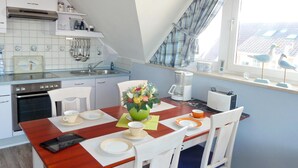 Een eetgedeelte met een houten tafel, witte stoelen en een bloevas. Achtergrond is een keuken met een fornuis, spoelbak en keukengerei.