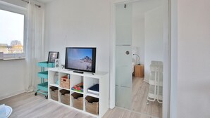 Un soggiorno moderno con una televisione su un mobile bianco, un carrello portaoggetti color petrolio e una porta vetrata che dà su un bagno.