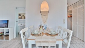 Zona pranzo con tavolo bianco apparecchiata per due, sottopiattino in tessuto intrecciato e lampada a sospensione.