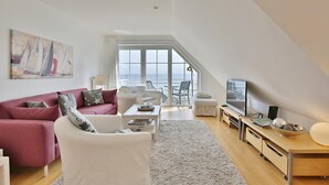 Un salon moderne avec un canapé rouge, un fauteuil blanc et une table basse. On y trouve un téléviseur posé sur un meuble en bois, ainsi qu’une grande fenêtre donnant sur la mer.