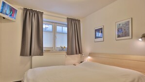 Ett hotellrum med en stor säng, ett fönster med gardiner, ett element, en tv på väggen och två inramade tavlor.