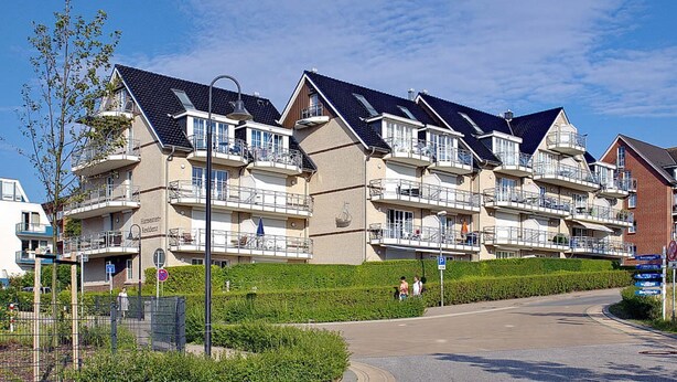 Una hilera de edificios residenciales modernos de varios pisos, con balcones, un cielo despejado y una zona verde bien cuidada al frente.