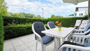 Un patio al aire libre con una mesa y sillas blancas, una pequeña planta en maceta y un seto de fondo.