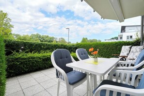 An outdoor patio with a white table and chairs, a small potted plant, and a hedge in the background. - Hanseaten-residenz Appartement 6 (Scharbeutz)