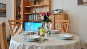 Una mesa de comedor preparada con platos, cubiertos y copas de vino, rodeada de sillas, con una estantería y una pequeña planta al fondo.