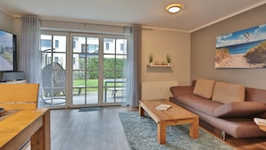 Un salon moderne avec un canapé, une table basse et un téléviseur posé sur un meuble en bois. Une porte-fenêtre coulissante donne accès à un patio équipé d’une table et de chaises.