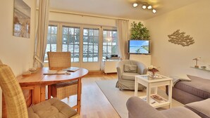A cozy living room with a wooden dining table, comfortable seating, a television, and a large window with curtains. - Seepark Appartement 13 (Scharbeutz)
