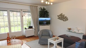 A living room with a television, a sofa, a chair, a small table, a radiator, and a window with curtains. - Seepark Appartement 13 (Scharbeutz)