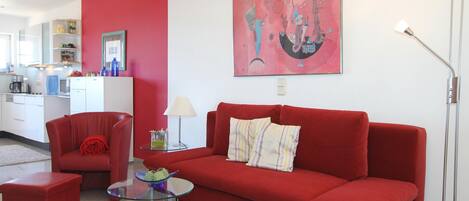 A modern living room with a red sofa, a glass coffee table, and a red armchair. A kitchen is visible in the background.