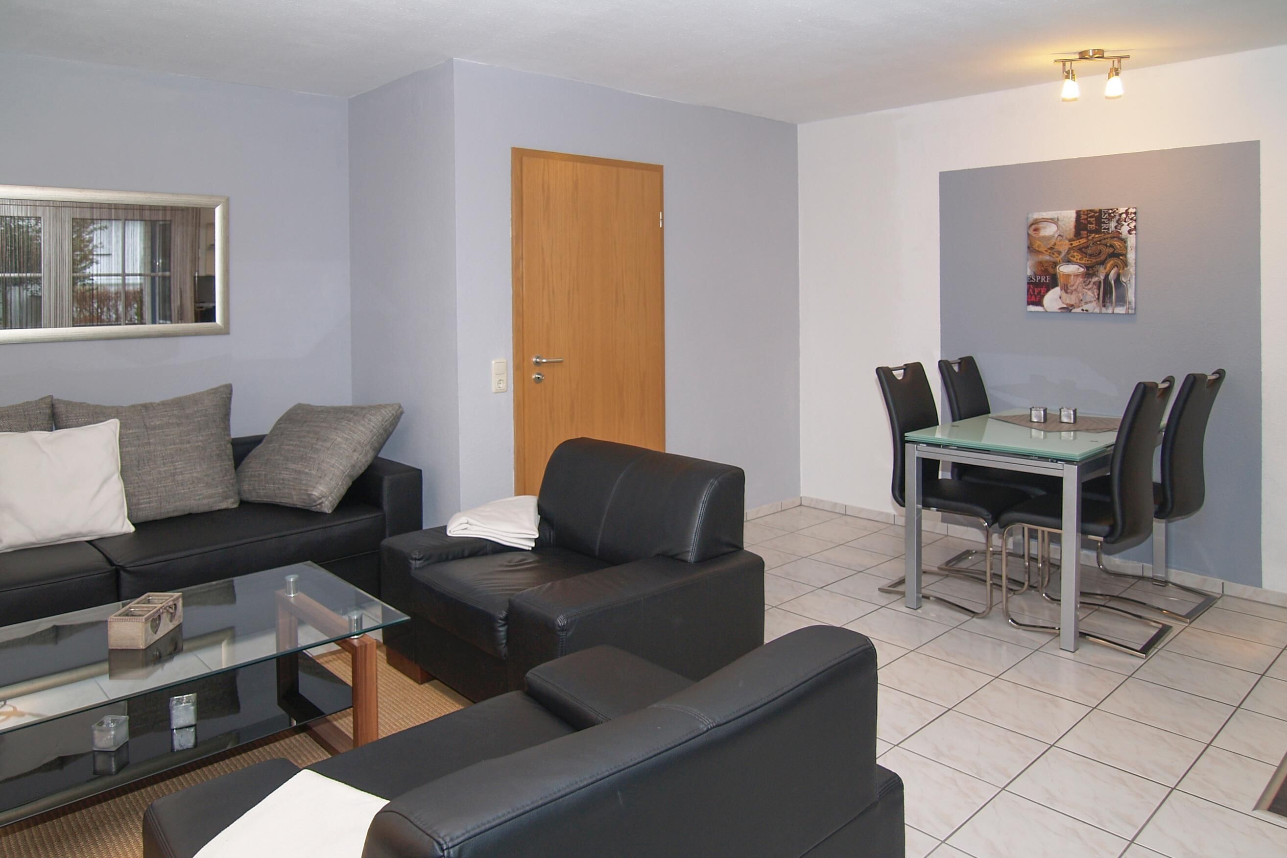 A modern living room with a glass coffee table, black leather sofas, a dining table with chairs, and a wooden door.