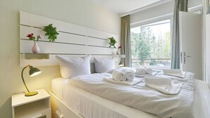 A hotel room with a neatly made bed, a bedside lamp, a small shelf with a plant, and a window with curtains. - Residenz am Hang Appartement 103 (Scharbeutz)