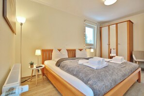 Un lit bien fait, recouvert d’une couverture grise, une lampe de chevet et une petite table sur laquelle est posée une plante.