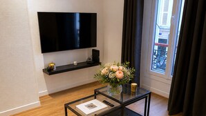 A modern living room with a flat-screen TV, a glass coffee table with books, a vase with flowers, and a window with curtains.