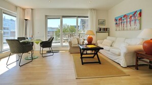A modern living room with a white sofa, a dining table with chairs, a coffee table, and a painting on the wall. - Barke Appartement 324 (Scharbeutz)