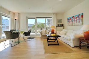 A modern living room with a white sofa, a dining table with chairs, a coffee table, and a painting on the wall. - Barke Appartement 324 (Scharbeutz)