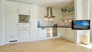 A modern kitchen with white cabinetry, stainless steel appliances, a flat-screen TV, and a trash can. - Barke Appartement 324 (Scharbeutz)
