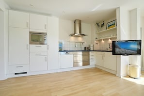 A modern kitchen with white cabinetry, stainless steel appliances, a flat-screen TV, and a trash can. - Barke Appartement 324 (Scharbeutz)