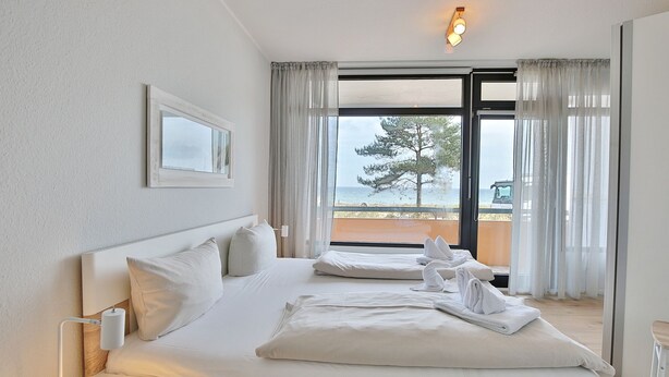 A hotel room with a large bed, a bedside table, a nightstand, and a view of the sea through a sliding glass door. - Berliner Hof Appartement 116 (Scharbeutz)