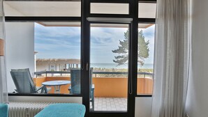 A room with a large sliding glass door leading to an outdoor balcony with a view of the sea and a thatched-roof structure. - Berliner Hof Appartement 116 (Scharbeutz)