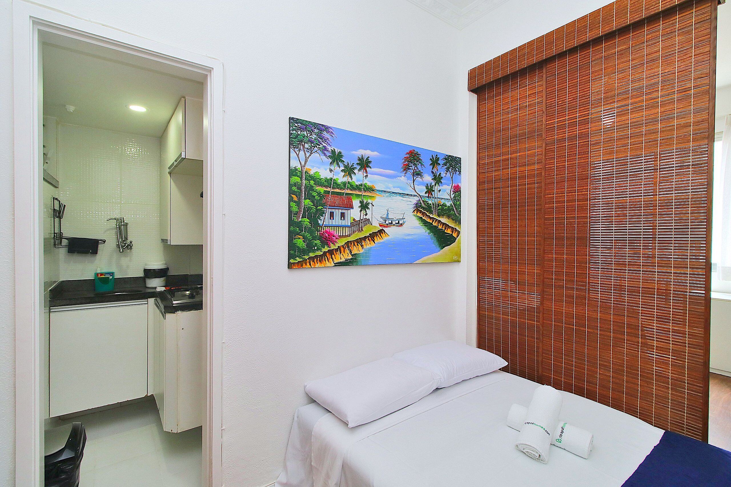 Hotelkamer met een bed, een schilderij van een tropisch landschap, een houten zonnescherm en een badkamer die door een open deur zichtbaar is.