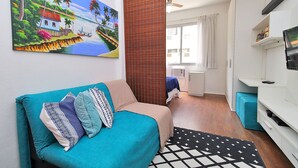 A modern living room with a teal sofa, a painting of a tropical scene, a wooden partition, and a white entertainment unit. - Rio Spot Homes C039 (Rio de Janeiro)