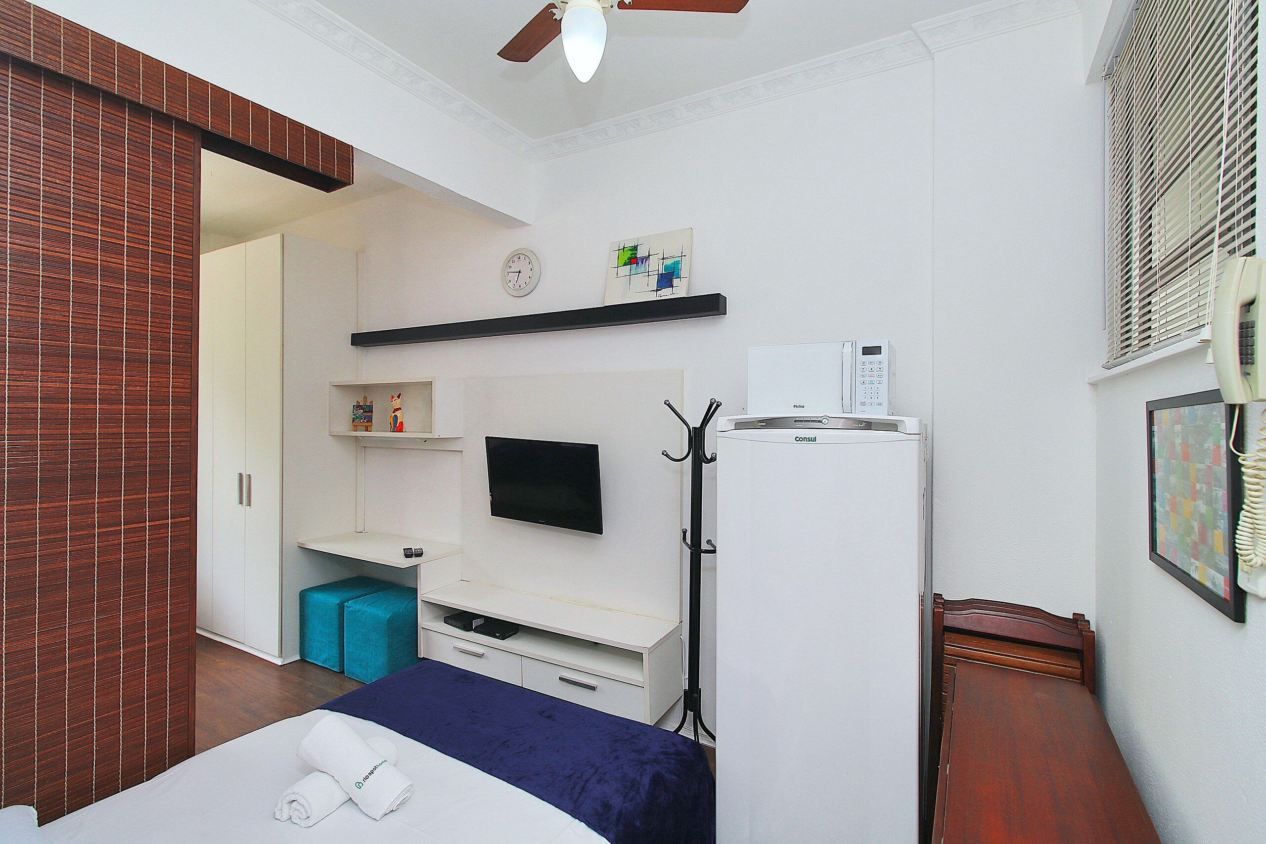 Habitación de hotel con cama, escritorio, televisión, refrigerador y ventana con persianas.