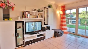 Ein modernes Wohnzimmer mit Fernseher, Bücherregalen, einem Schrank und einem großen Fenster mit Vorhängen.
