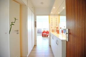 A modern kitchen with white cabinets, a wooden door, and a view into a living area with a red sofa. - Perle de la Plage Apartment 403 (Haffkrug)