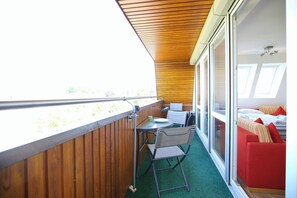 A balcony with a wooden ceiling, a small table, and a chair. There is a bed with red and white bedding in the background.