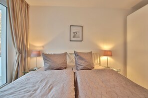 A double bed with a geometric patterned bedspread, two bedside lamps, a framed picture on the wall, and a window with curtains. - Strandglück Appartement 12A (Scharbeutz)