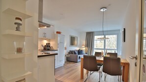 A modern kitchen with white cabinets, a dining area with a wooden table and chairs, and a living area with a sofa and a large window.