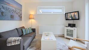 Una sala moderna con un sofá gris, un cojín a rayas, una lámpara de pie, una mesa de centro blanca, un televisor y una ventana con vista a la playa.