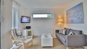 A modern living room with a grey sofa, a wooden chair, a white coffee table, a TV mounted on the wall, and a window with a view of the outdoors. - Haus Küstenkinder Louis Appartement 2 (Scharbeutz)