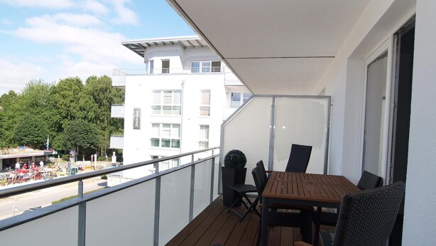 A balcony with a wooden table and chairs, a glass railing, and a view of a street with buildings and trees. - Strandglück Appartement 12B (Scharbeutz)