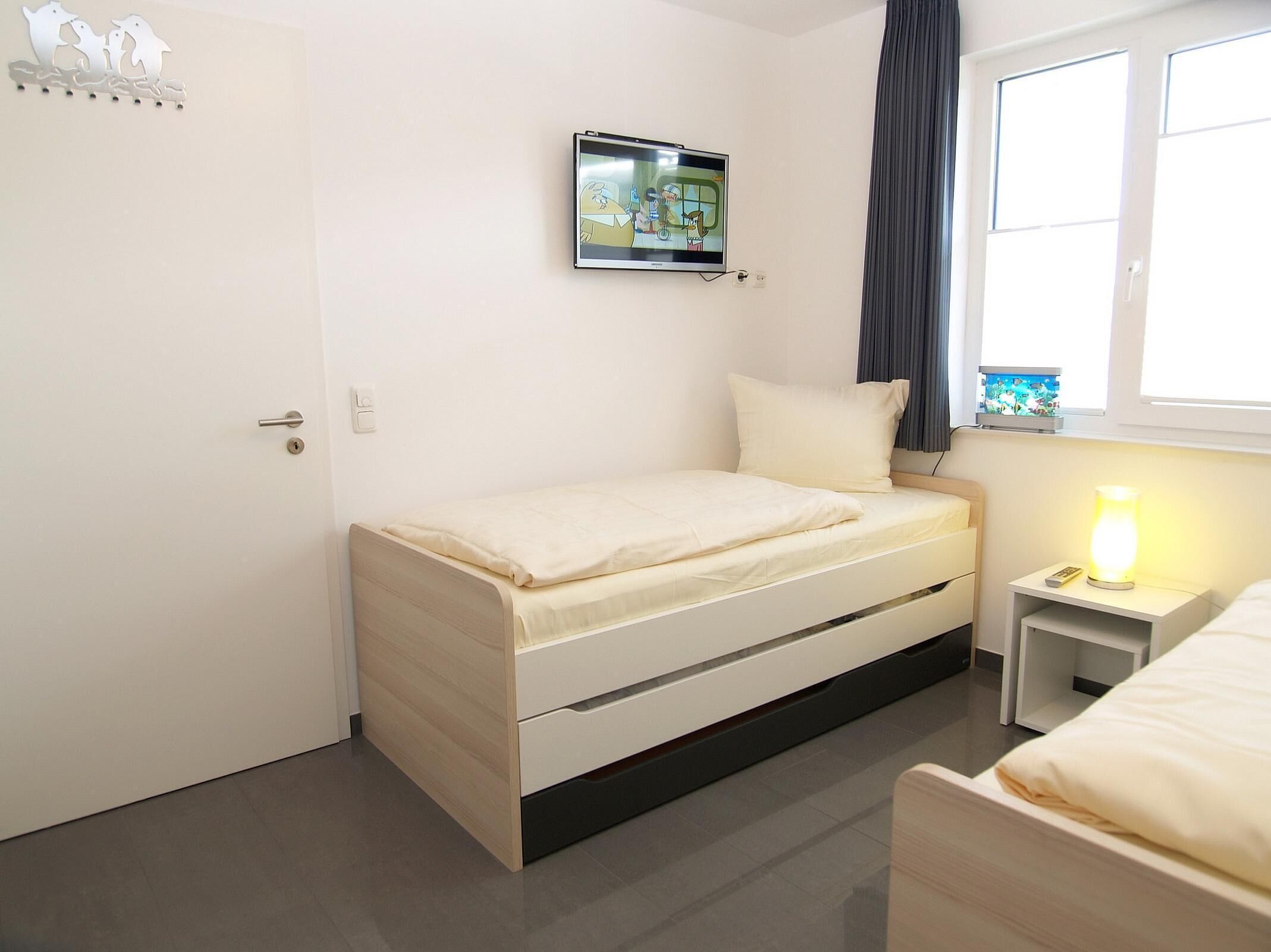 Une petite chambre à coucher, bien rangée, avec un lit simple, une table de chevet avec une lampe et un téléviseur fixé au mur.