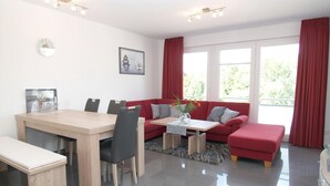 A modern living room with a dining table, red sofa, and a large window with curtains. - Strandglück Appartement 12B (Scharbeutz)