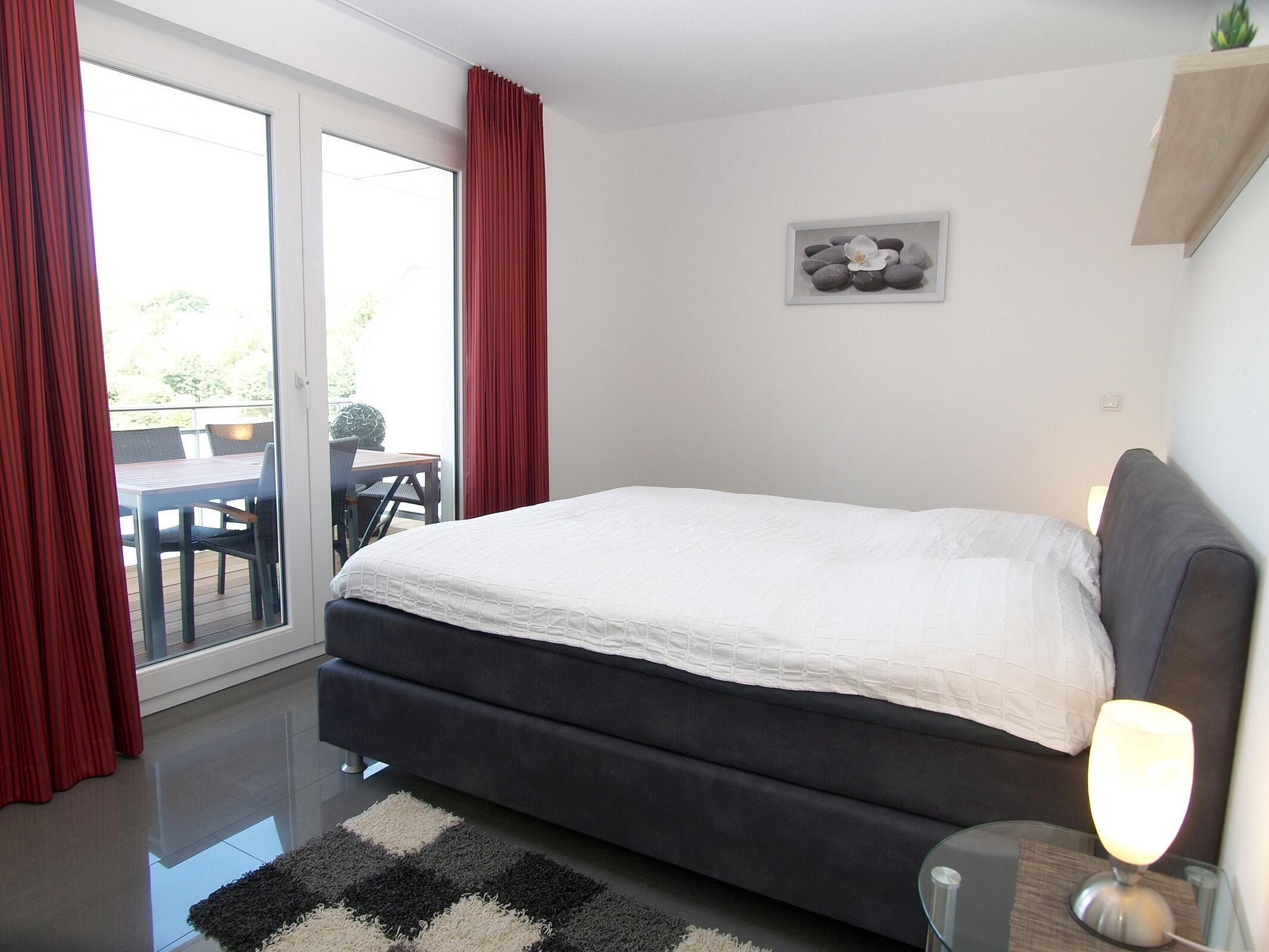 Une chambre moderne avec un grand lit, une porte vitrée donnant sur un balcon et une petite table avec une lampe.