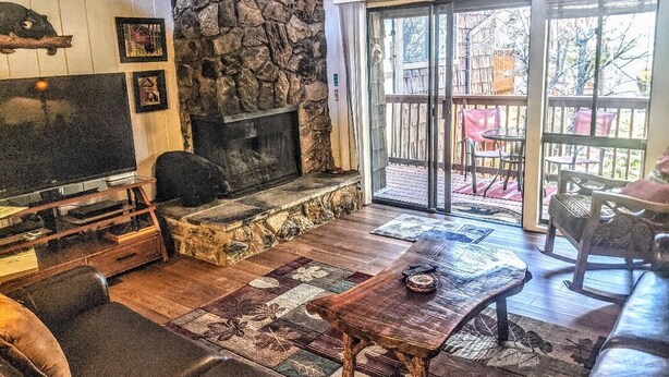 A cozy living room with a stone fireplace, a television, and a large window overlooking a deck. - Ski, Soak & Relax, 2 BR/1.5 BA Townhouse with Spa, at the base of Snow Summit (Big Bear Lake)