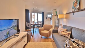 Ein modernes Wohnzimmer mit Fernseher, Sofa, Sessel, Essbereich und Küche.