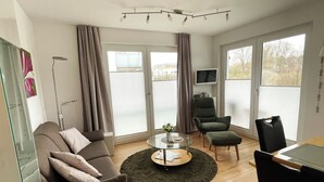 A modern living room with a sofa, armchair, ottoman, and a glass coffee table. There is a TV mounted on the wall and a window with blinds. - Strandglück Appartement 9B (Scharbeutz)