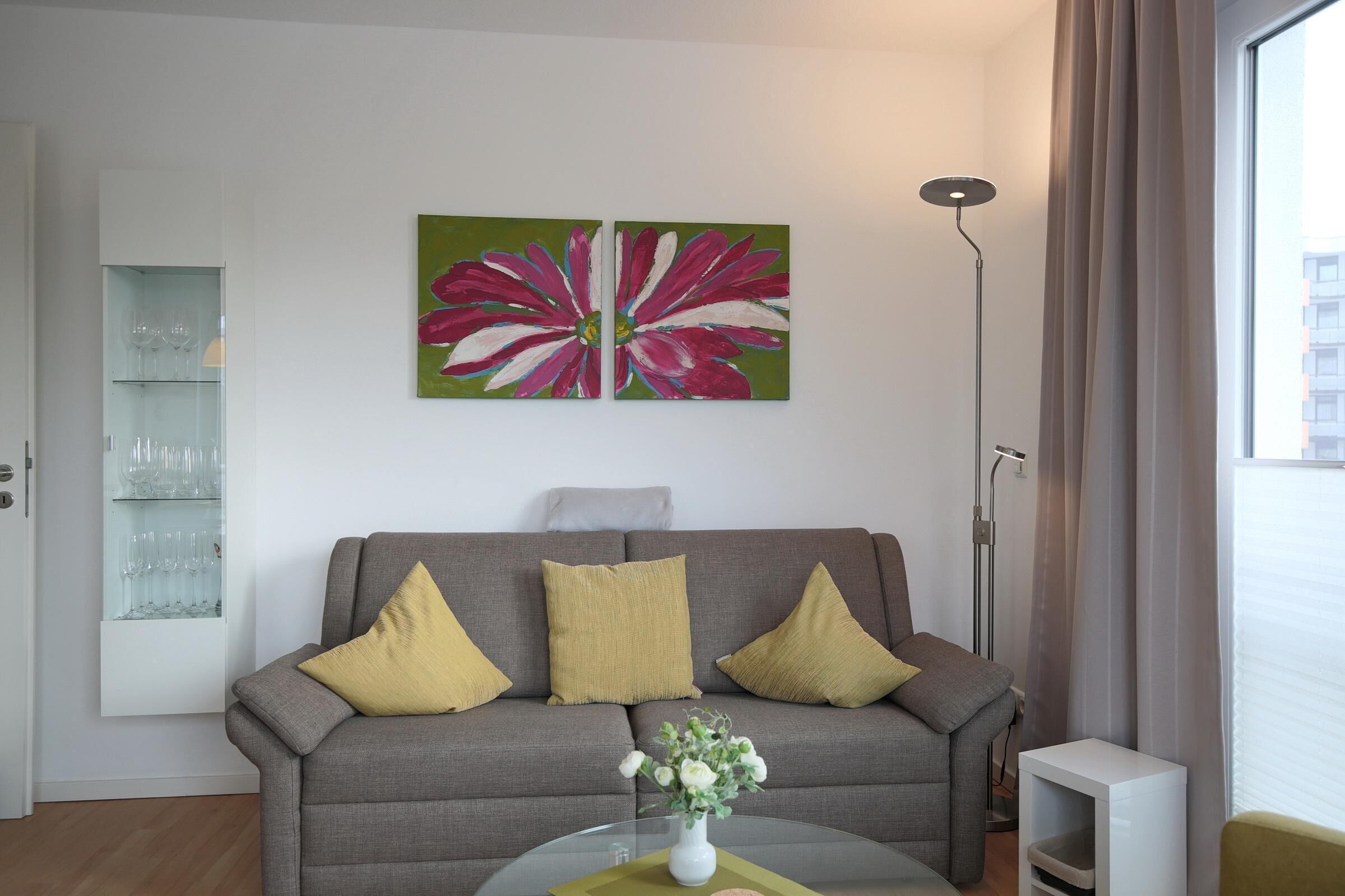 A modern living room with a grey sofa, yellow cushions, a glass coffee table with a vase of flowers, and a large floral painting on the wall.