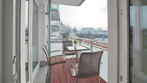 A balcony with a glass railing, wicker chairs, a small table with a vase, and a view of buildings in the distance. - Strandglück Appartement 9B (Scharbeutz)