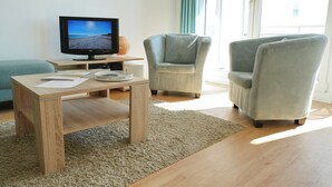 Una sala de estar con una mesa de centro de madera, dos sillones, un televisor y un ventanal con cortinas.