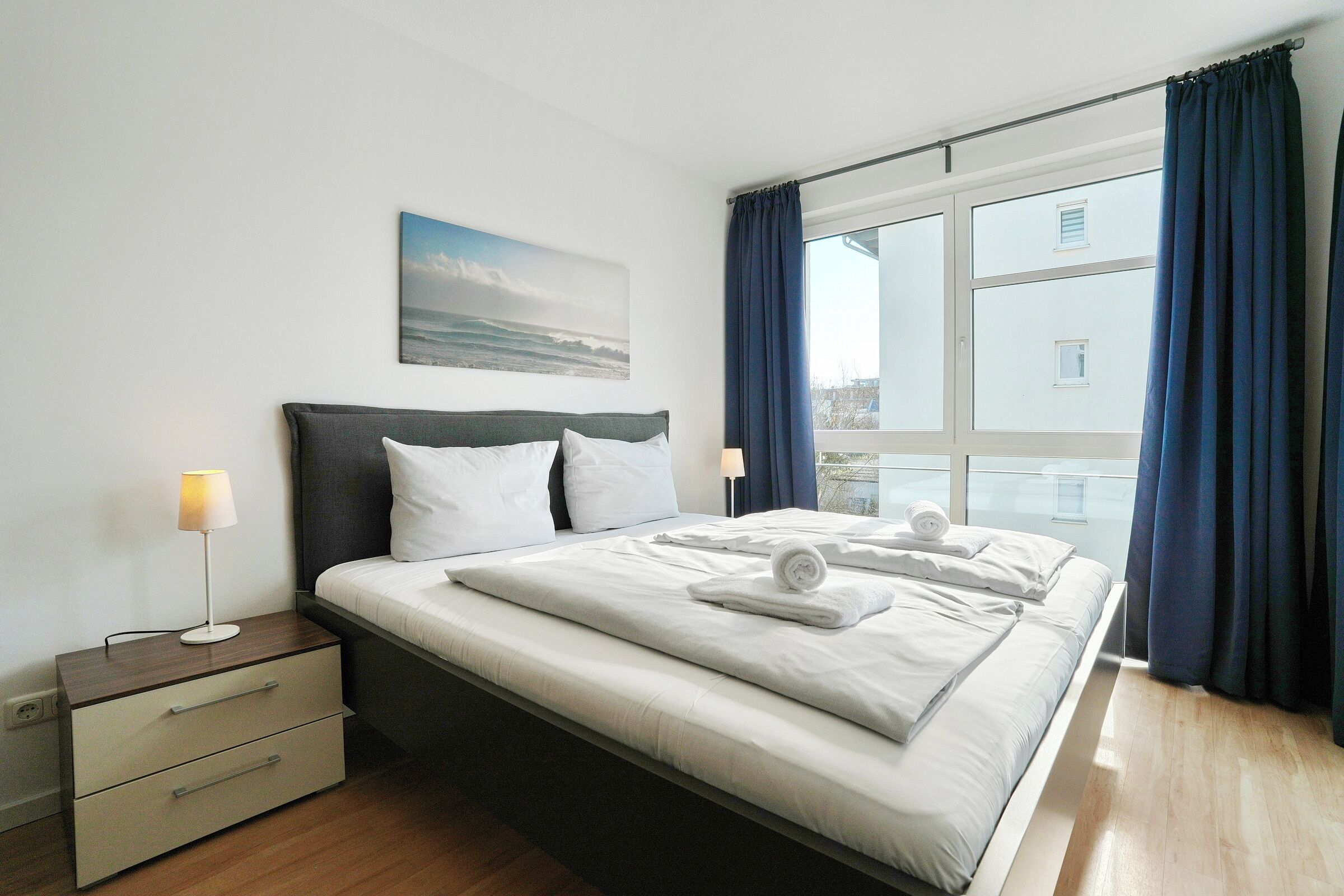 Quarto de hotel moderno com uma cama grande, uma mesa de cabeceira, uma luminária e vista para a cidade através de uma janela.